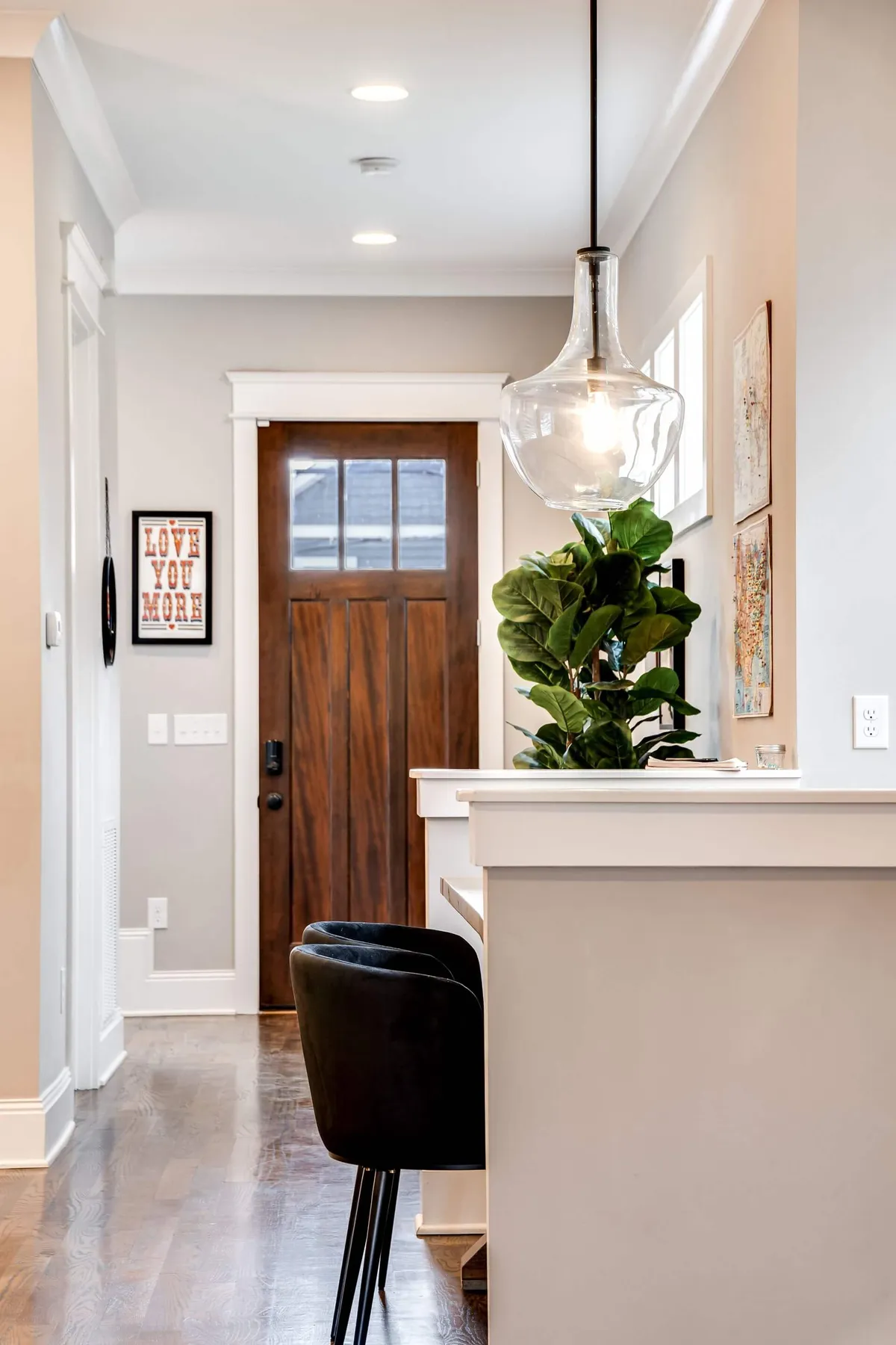 The image features a bright, inviting entryway with the front door, a hanging light fixture, bar stools facing an open kitchen area, a decorative plant, and wall art. The space feels welcoming and sets the tone for the rest of the home.