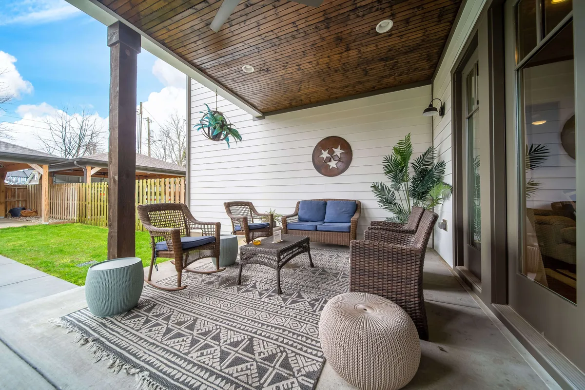 A stylish backyard patio with wicker seating arranged around a coffee table, accented by a decorative rug and hanging plant. The area is complemented by wall decor, creating a cozy, welcoming space perfect for gatherings and relaxation.