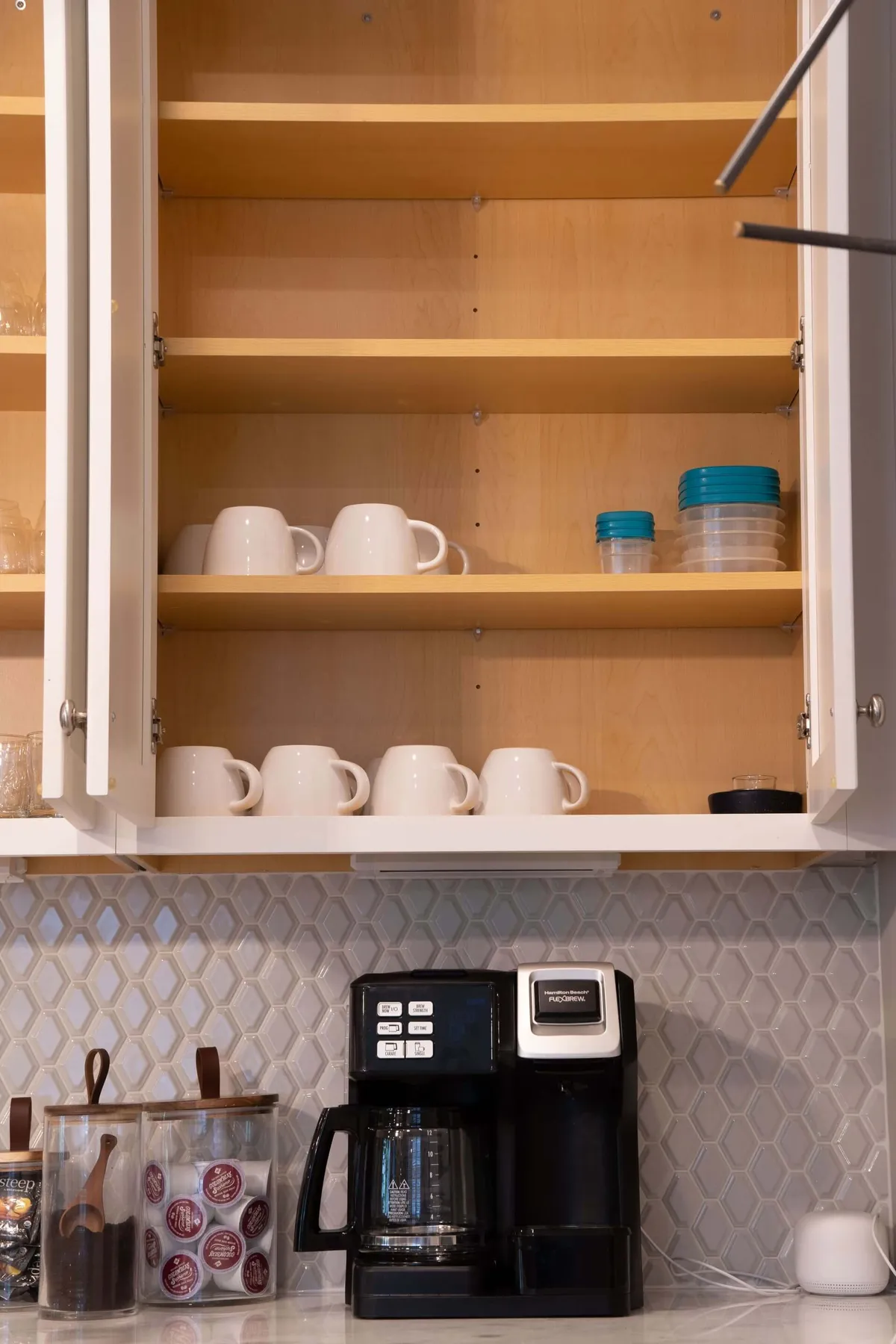 A close-up of a coffee station featuring a coffee maker, white mugs, storage containers, and a hexagonal tile backsplash, evoking cozy, inviting mornings and snack breaks.