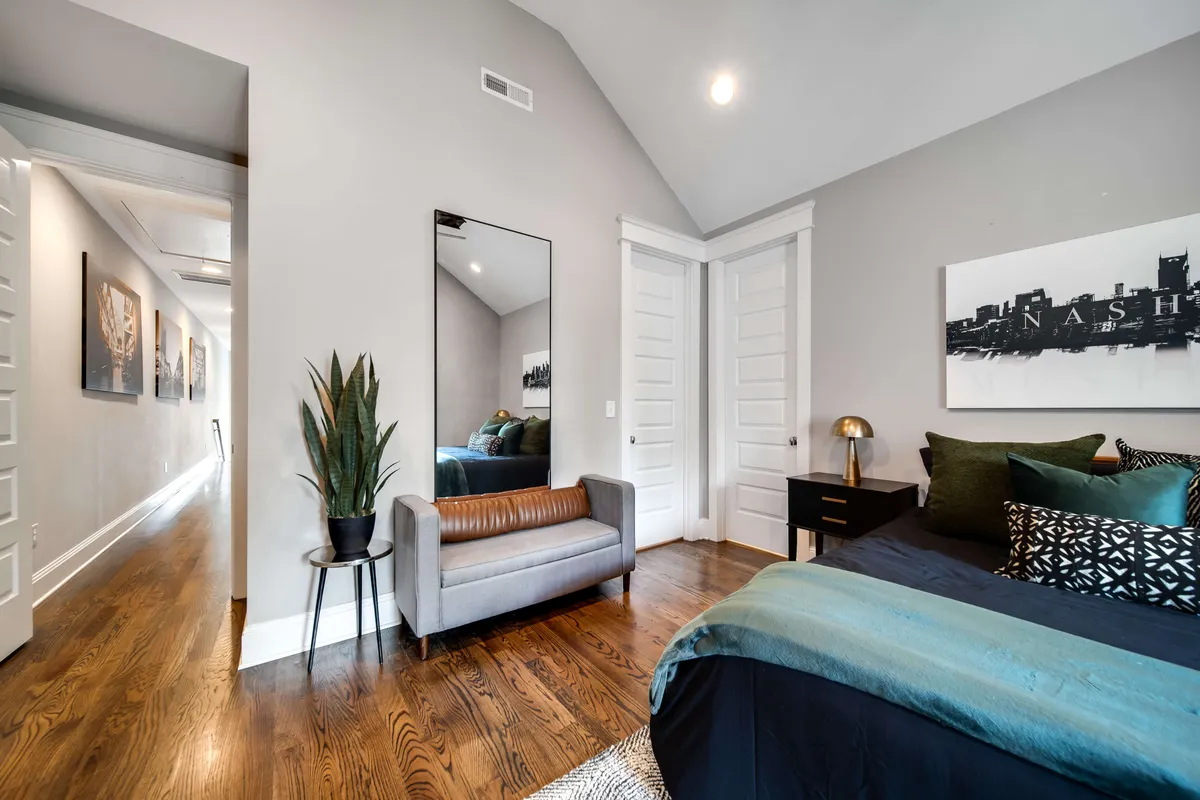 This stylish bedroom features a bed with decorative pillows, a full-length mirror, a contemporary art piece on the wall, a potted plant, and an accent chair. The room feels inviting and modern, offering both relaxation and space for socializing or preparing for the day.