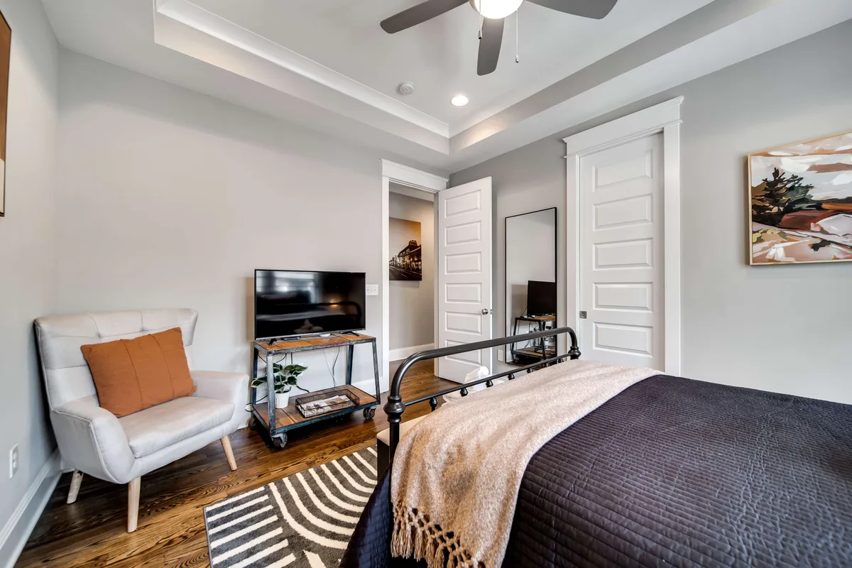 A cozy bedroom scene with a bed dressed in dark bedding, a comfortable armchair with an orange cushion, a television and stand positioned conveniently for in-bed viewing, and vibrant artwork on the wall. There is a sense of modern comfort and personal space.