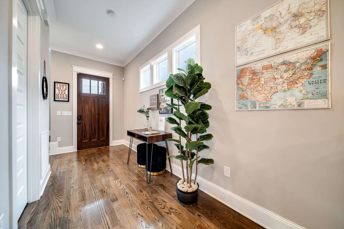 This inviting entryway features a wooden front door, a decorated console table, a potted plant, and map artwork on the wall. Natural light brightens the space, creating a warm first impression and a sense of connection between the home and the city outside.