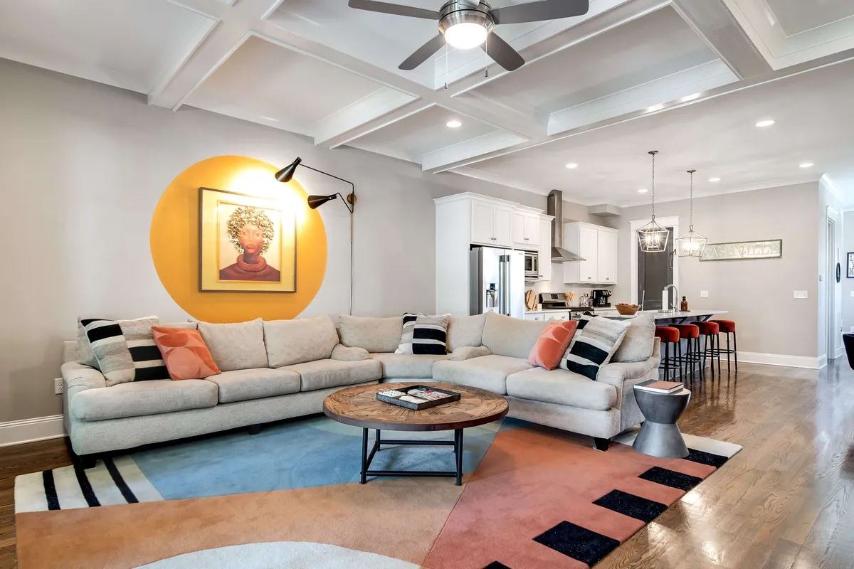 A spacious living room featuring a large sectional sofa, a colorful area rug, framed artwork, and pendant lighting. The arrangement is inviting and perfect for group gatherings, with ample soft seating and stylish decor that blends industrial and southern influences.