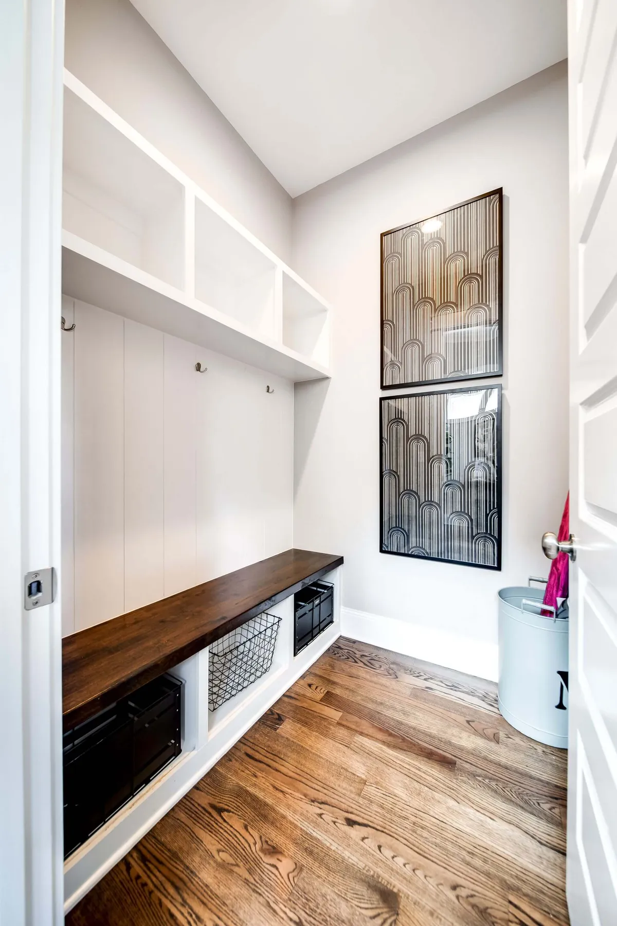 A functional mudroom scene with a wooden bench, open shelving holding storage baskets, and decorative wall art. The space is tidy and organized, designed for storing shoes, coats, and bags after a busy day out.