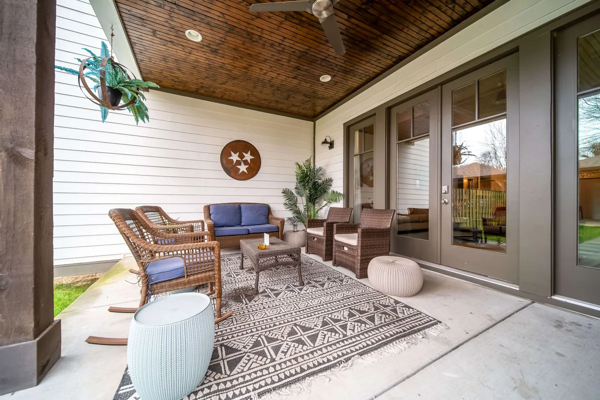 An inviting outdoor scene featuring woven seating and a coffee table set atop a decorative rug, with lush plant accents and double doors leading inside. The space feels tranquil and versatile, suitable for socializing, enjoying drinks, or reading.