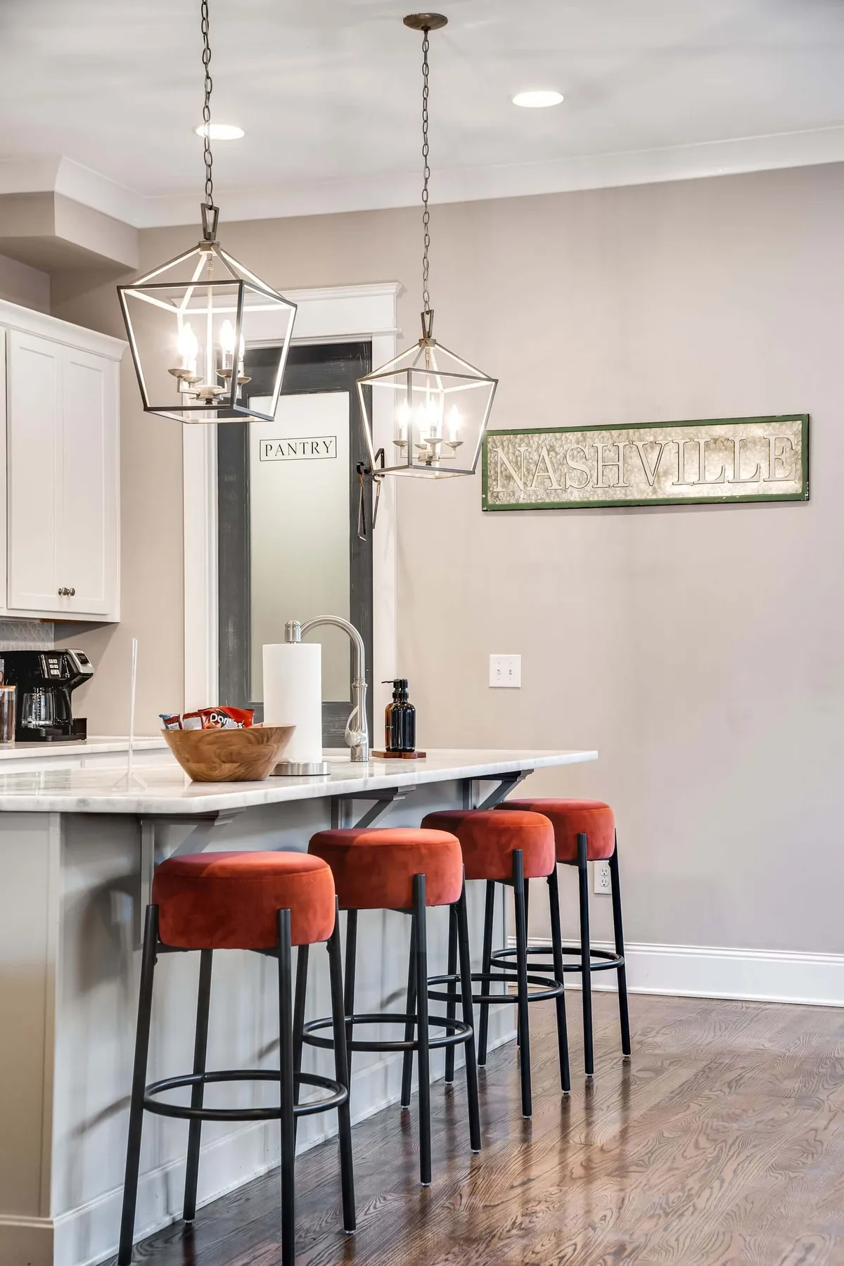 A modern kitchen with an island and stools, pendant lights, a pantry door, and a Nashville sign in the background. The space is designed for cooking, entertaining, and casual family dining, blending modern comfort with local flair.