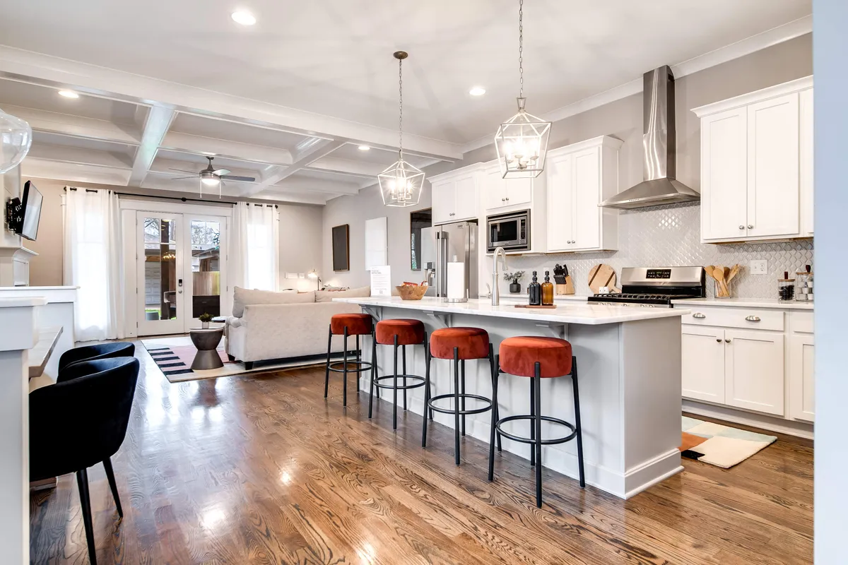 An open-plan kitchen and living area with a kitchen island, bar stools, white cabinetry, modern appliances, and a view of a spacious living area. The setup encourages gathering, socializing, and celebrating together before or after game day outings.